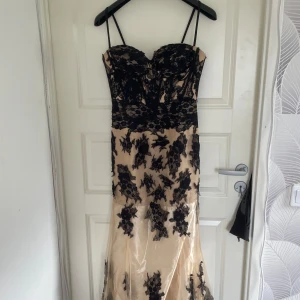 Svart spetsklänning med nude underklänning från Luxstore - Säljer en elegant långklänning med nude bas och svart spetsdetaljer. Klänningen har smala axelband, markerad byst och vacker spets över hela plagget. Perfekt för bal eller festliga tillfällen. Aldrig använd