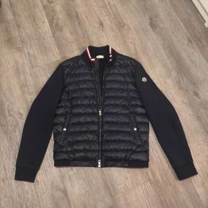 Moncler cardigan mörkblå - Snygg mörkblå cardigan från Moncler, Jackan har dragkedja, två sidofickor med knappar och en krage med röd, vit och blå rand. Varan är äkta och kan äkthets verifieras på monclera hemsidan, varan är köpt ifrån luxuriosecret på Instagram. Det finns en reva se bild 4. Inget som syns!