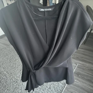 Svart omlottopp från Zara - Snygg svart omlottopp från Zara med v-ringning och knyt i sidan. Toppens material känns mjukt och har en elegant drapering framtill. Perfekt för dig som vill ha en stilren och trendig look.