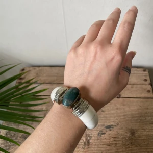 Brett armband med vit bas och grön mossagat - Unikt armband med bred vit bas och stora silverfärgade detaljer. I mitten sitter en stor oval grön sten som ger ett coolt statement. Armbandet har en rustik känsla och är perfekt för dig som gillar att sticka ut. Gjort i trä, horn, metall och mossagat.