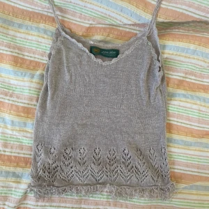 Beige stickad linnetopp från Jin Xin Products - Säljer en beige stickad linnetopp med smala axelband och spetsdetaljer längs halsringningen. Toppen har ett fint mönster nertill och fransar längs nederkanten. Perfekt för varma dagar och somriga outfits.