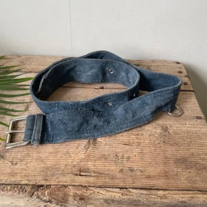 Blått mockabälte med silverfärgat spänne - Säljer ett brett blått bälte i mocka med silverfärgat metallspänne och extra ringdetaljer. Bältet har flera hål för justering och en mjuk, matt yta. Perfekt för att ge outfiten en cool touch.