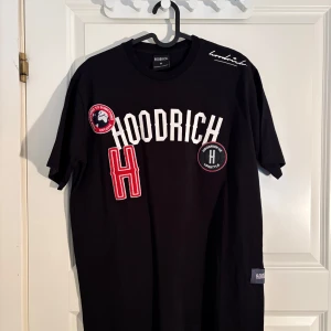 Svart t-shirt från Hoodrich med tryck - Säljer en svart t-shirt från Hoodrich i storlek M. T-shirten har flera tryck och broderade detaljer framtill, bland annat stora bokstäver och logotyper. Rund halsringning och korta ärmar. Perfekt för en streetwear-look.