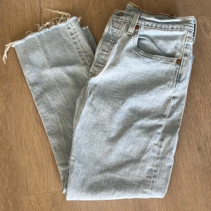 Ljusblå Levi's 501 jeans med råa benslut - Säljer ett par klassiska ljusblå Levi's 501 jeans med raka ben och råa, fransiga benslut. Jeansen har fem fickor, knappgylf och den ikoniska läderpatchen bak i midjan. Perfekta för en avslappnad och trendig look. Storlek  17-26