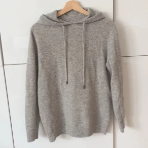  stickad hoodie - Mysig ljusgrå stickad hoodie med huva och dragsnören. Tröjan har lång ärm och ribbade muddar vid ärmslut och nederkant. Perfekt för kyliga dagar och enkel att matcha med olika outfits.