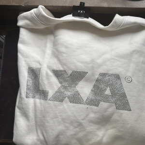 Vit sweatshirt från LXA med strasslogga - Säljer en vit sweatshirt från LXA med stor logga framtill i strass. Tröjan har rund halsringning och långa ärmar. Perfekt för en stilren och trendig look.( Tvättad en gång. En pytteliten svart fläck på ryggen och några typ 10 små stenar som trillat av. Annars är den verkligen i nyskick). Skriv för bilder. 