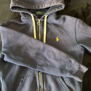 Blå hoodie från Polo Ralph Lauren med gul dragkedja - Snygg blå  hoodie från Polo Ralph Lauren med gul dragkedja och matchande gula snören. Klassisk gul logga broderad på bröstet. Mjuk insida och två fickor framtill. Perfekt för en avslappnad stil.