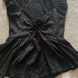 Svart topp med volang från 157 - Svart topp från 157 i storlek XS med volangdetaljer vid axlarna och en snygg knytdetalj framtill. Toppen har korta ärmar och ett lätt glansigt tyg som ger en elegant känsla. Perfekt för att lyfta din outfit.