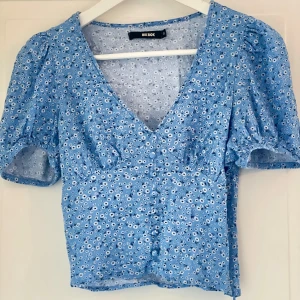 Blommig blå topp från Bik Bok - Supersöt blå topp med små vita blommor från Bik Bok. Blusen har v-ringning, kort puffärm och knappar framtill. I mycket bra skick!