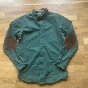 Ralph Lauren skjorta  - Säljer en grön långärmad skjorta från Polo Ralph Lauren med bruna mockadetaljer på armbågarna och klassisk krage. Skjortan har knappar framtill och en liten broderad logga på bröstet. Perfekt för en stilren och avslappnad look. Har en liten vit fläck som syns på bild 3 ;)