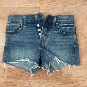 Levi's jeansshorts - Säljer ett par klassiska blå jeansshorts från Levi's med råa fransade kanter och knappar framtill. Shortsen har fem fickor och en hög midja. Perfekta för sommaren och ger en avslappnad vintagekänsla. I storlek 25! Pris går att diskutera💕