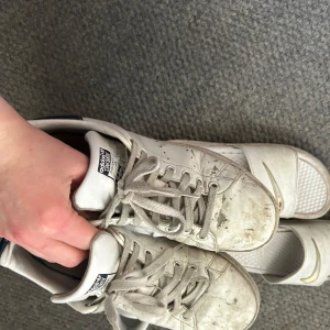 Vita Adidas Stan Smith sneakers - Klassiska vita Adidas Stan Smith sneakers med gröna detaljer på hälen. Skorna har snörning och platt sula, samt rund tå. Perfekta för en avslappnad och sportig stil.