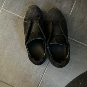 Svarta sneakers från Axel Arigato - Svarta sneakers från Axel Arigato med minimalistisk design och elastiskt band över vristen istället för klassisk snörning. Skorna har rund tå och platt sula, tillverkade i skinn och syntetmaterial. Perfekta för en clean och stilren look.