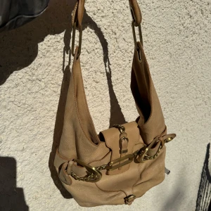 Beige axelväska från Jimmy Choo med gulddetaljer - Klassisk Jimmy Choo axelväska med snygga guldfärgade metalldetaljer och kedja. Väskan är tillverkad i skinn och har ett innerfack med dragkedja. Perfekt för dig som vill ha en stilren och lyxig accessoar.