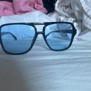 Svarta solglasögon med blåtonade glas - Snygga solglasögon med svart båge och blåtonade, fyrkantiga glas. Bågen har en cool detalj i guld upptill vid näsryggen. Perfekt accessoar för att lyfta din stil under soliga dagar.
