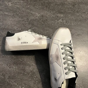 Golden Goose skor med grå stjärna  - Säljer ett par vita sneakers från Golden Goose med grå snörning och en grå stjärna på sidan. Skorna har svart hälparti och klassisk platt sula. Perfekta för dig som gillar stilrena och trendiga sneakers.