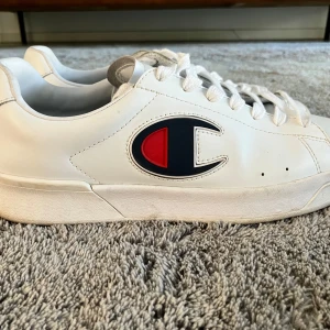 Vita sneakers från Champion - Säljer ett par vita sneakers från Champion med klassisk logga i rött och blått på sidan. Skorna är robusta och lite tjockare i material, har snörning, rund tå och platt sula. Perfekta för en clean och sportig stil.