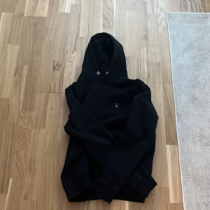 Svart hoodie från Gant - Svart hoodie från Gant, har en enkel design med diskret logga på bröstet och ribbade muddar vid ärmslut och nederkant. Perfekt för en avslappnad stil.