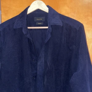 Mörkblå manchesterskjorta från Massimo Dutti - Säljer en stilren mörkblå skjorta i manchester från Massimo Dutti. Skjortan har klassisk krage, knäppning framtill och långa ärmar. Perfekt för dig som vill ha en tidlös och snygg skjorta i garderoben.