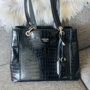 Svart handväska i croco-mönstrat skinn från Guess - Säljer en svart handväska från Guess med croco-mönstrad yta och silverfärgade metalldetaljer. Väskan har dubbla handtag och dekorativa tags. Perfekt för dig som vill ha en stilren och trendig accessoar.