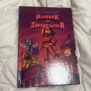 Handbok för superhjältar - En spännande barnbok med röd och lila hård pärm, skriven av Elias & Agnes Våhlund. Omslaget visar flera superhjältar i färgglada dräkter på ett tak framför en klocka och kupol. Perfekt för unga läsare som gillar äventyr och superkrafter.
