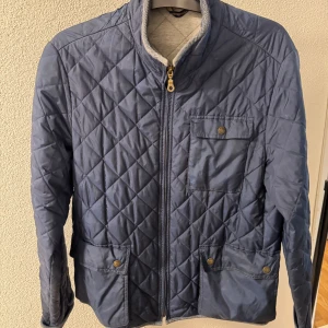 Blå quiltad jacka från Henri Lloyd - Säljer en blå quiltad jacka från Henri Lloyd med dragkedja och tryckknappar på fickorna. Jackan har lång ärm och ett broderat märke på ena ärmen. Perfekt för höst och vår, stilren och klassisk design.