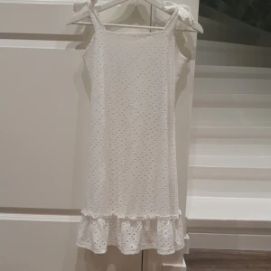 Vit klänning med rosetter och en volangkant - Söt vit klänning med knytbara axelband och broderat hålmönster. Klänningen har en volangkant nertill och är ärmlös, perfekt för varma dagar. Lätt och luftig modell. Fin till skolavslutning eller studentfest