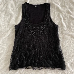 Svart spetslinne med pärldetaljer - Säljer ett svart linne från pimkie med vacker spets och pärldetaljer framtill. Linnet är ärmlöst och har rund halsringning. Perfekt för dig som vill ha något extra till outfiten!