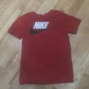 Röd Nike t-shirt med logga - Röd t-shirt från Nike med stor vit och svart Nike-logga på bröstet. Klassisk passform och rund halsringning. Perfekt för en sportig och avslappnad stil.