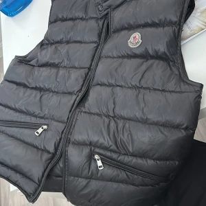 Svart dunväst från Moncler - Säljer en svart dunväst från Moncler med quiltad design och dragkedja framtill. Västen har två fickor med dragkedja och en liten Moncler-logga på bröstet. Perfekt för lager-på-lager-stil.