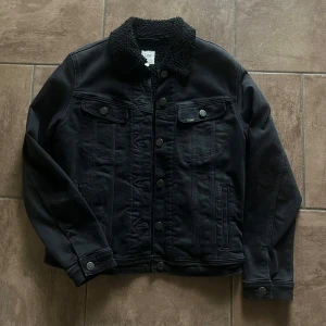 Fin svart jeansjacka från Lee - Säljer en svart jeansjacka från Lee med mysigt teddyfoder och krage. Jackan har knappar framtill, två bröstfickor med lock och knapp samt sidofickor. Baktill finns två knappar för att justera midjans passform. Storlek S, unisex modell.