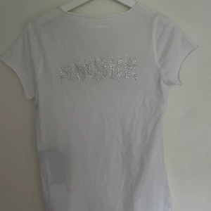 Vit t-shirt med strassdetaljer från Zadig & Voltaire - Vit t-shirt med v-ringning och korta ärmar från Zadig & Voltaire. Baksidan har texten 'AMOUR' i glittrande strass. Tröjan är helt oanvänd med prislappen kvar, men hr ett litet hål i sidan (se bild 4). Nypris: 1300kr