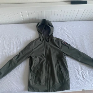 Olivgrön softshelljacka med huva från Parajumpers - Säljer en olivgrön softshelljacka från Parajumpers med huva och dragkedja. Jackan har två snedställda fickor framtill och detaljer i orange på insidan. Perfekt för dig som vill ha en stilren och funktionell jacka.