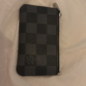 Rutig plånbok  - Säljer en stilren liten plånbok  svart och grått med klassiskt rutigt mönster. Plånboken har dragkedja upptill och är perfekt för kort och mynt. Snygg accessoar att ha med sig i fickan eller väskan.