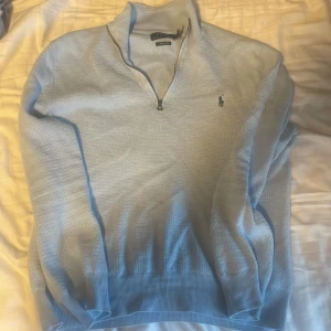 Ljusblå halvzip tröja från Polo Ralph Lauren - Säljer en ljusblå tröja från Polo Ralph Lauren med halv dragkedja och klassisk krage. Tröjan har lång ärm och diskret broderad logga på bröstet. Perfekt för lager-på-lager och stilrena outfits.