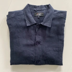 Mörkblå overshirt i linne från SIR - Stilren mörkblå overshirt i linne med klassisk krage och tre fickor framtill. Skjortan har knäppning med matchande knappar och lång ärm. Perfekt för en avslappnad men snygg look. nypris 2000kr.
