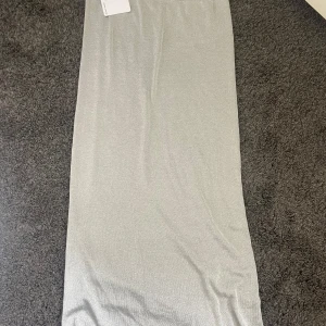 Silverglittrig långkjol - Säljer en snygg långkjol i silver med glittrigt tyg. Kjolen har en rak passform och är perfekt för dig som vill sticka ut med en stilren look. Passar till många olika tillfällen när du vill glänsa lite extra.