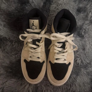 Nike Air Jordans i beige och svart - Säljer ett par Nike Air Jordans med beige mockadetaljer och svarta partier. Skorna har klassisk snörning, hög modell och den ikoniska swoosh-loggan på sidan. Perfekta för dig som gillar streetwear och sneakers med stil. Storlek 38