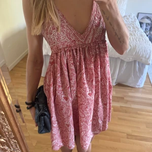 Röd/vit/rosa kortklänning med paisleymönster - Säljer en röd och vit/ rosa kortklänning med paisleymönster och pärlor. Klänningen har v-ringning, är ärmlös och har öppen rygg med knytband i nacken. Modellen är luftig och har en omlottliknande överdel.