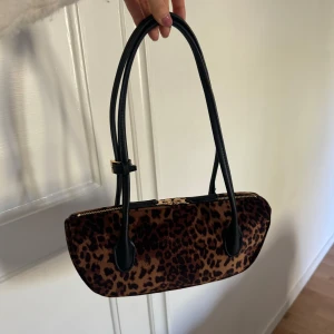 Leopardmönstrad axelväska - Säljer en snygg axelväska med leopardmönster, svarta handtag och guldiga detaljer. Väskan har dragkedja upptill och en avlång form. Perfekt för att lyfta vilken outfit som helst.