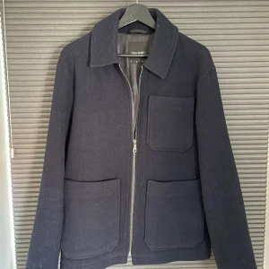 Mörkblå ulljacka från Lyle & Scott - Stilren mörkblå jacka från Lyle & Scott i ullblandning. Jackan har tre stora utanpåfickor, klassisk krage och dragkedja framtill. Ett lyle & scott märke ingår som aldrig använts. Köptes ny för 2000kr.