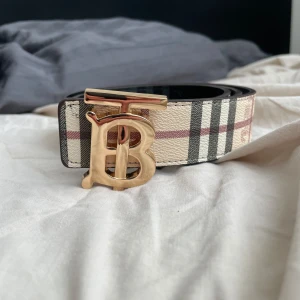 Burberry bälte med guldspänne - Säljer ett Burberry bälte med klassiskt rutigt mönster på ena sidan och svart på andra. Bältet har ett stort guldigt TB-spänne och är tillverkat i skinn. Perfekt accessoar för att lyfta din outfit.