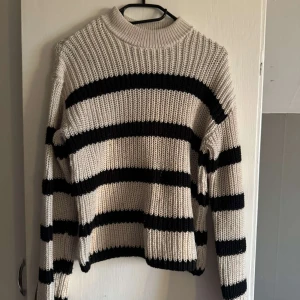 Randig stickad tröja med hög krage - Säljer en stickad tröja med breda svarta och beige ränder. Tröjan har lång ärm, ribbad struktur och en högre krage.💕