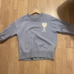 Ljusblå stickad tröja med hjärtdetalj - Säljer en ljusblå stickad tröja Ami Paris tröja med ribbade muddar och en vit hjärtdetalj med bokstaven A på bröstet. Tröjan har en avslappnad passform och är perfekt för kyligare dagar. Priset kan även diskuteras