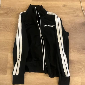Svart track jacket från Palm Angels - Säljer en svart track jacket från Palm Angels med vita ränder längs ärmarna och vit dragkedja. Jackan har hög krage och logga på bröstet. Perfekt för en sportig och trendig look.