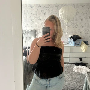 Svart tubtopp i satin - Säljer en svart tubtopp i glansigt material. Toppen har en lätt volang nertill och smockad resår i midjan. Perfekt att styla med jeans eller kjol. Perfekt till fest!!