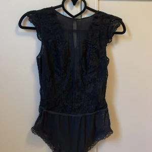 Svart spetsbody från H&M - Säljer en svart body från H&M med spetsdetaljer och korta ärmar. Bodyn har transparent spets framtill och baktill, samt fina volangdetaljer vid axlarna. 