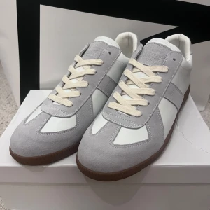 Maison Margiela  - Säljer ett par Maison Margiela skor i grå mocka och vitt skinn med vita snören, perfekta skor för sommaren. Är väldigt enkla att matcha med! Man får med lådan och resten av sakerna som är i. Storlek 42!