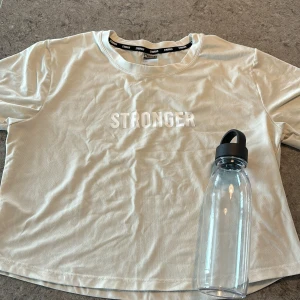 Beige croppad t-shirt från Stronger - Snygg beige croppad t-shirt från Stronger med vit text framtill. T-shirten har korta ärmar och är tillverkad i en mjuk blandning av återvunnen polyester och elastan. Perfekt för en sportig outfit till gymmet eller en avslappnad look!! Använd en gång. Väldigt bra skick! Pris diskuteras privat💕💕💕🫶🏼