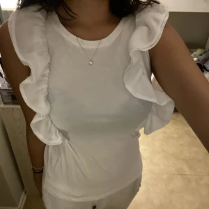 Vit ärmlös topp med volang - Säljer en vit ärmlös topp med breda volangdetaljer längs axlarna. Toppen har rund halsringning och är i ett mjukt, stretchigt material. Perfekt för en fräsch och stilren look.